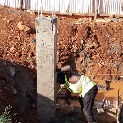 A construction worker in a bright yellow safety vest and black pants is working at a construction site in Borabu Chitago ward, Kenya. He is using a hammer or similar tool on the base of a tall, rectangular concrete column with exposed rebar extending from the top. The area around the column is excavated, revealing red-brown soil and rocks. There is standing water in the excavated area. A fence of corrugated metal sheets and wooden supports is visible in the background.