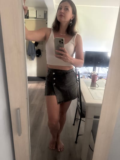 A woman is taking a mirror selfie in what appears to be an apartment. She is wearing a white tank top and a dark denim skirt. The background shows a room with a television, a table with a chair, and a mirror. There is also a closet area with clothing visible. The woman is barefoot. The location is the city of Saint-Claude, France.