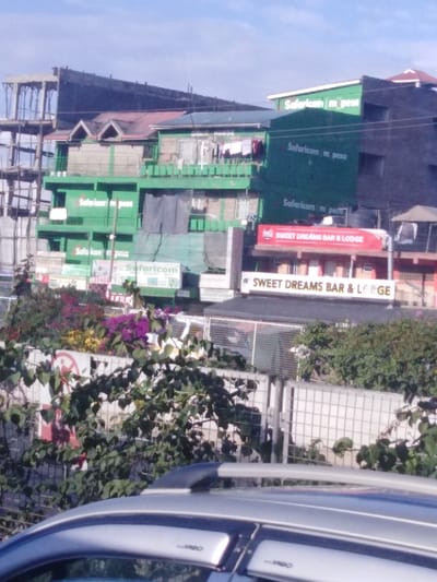The image shows several buildings with advertisements for "Safaricom m pesa" displayed on them. One building has a sign for "SWEET DREAMS BAR & LODGE," repeated on a lower level as "SWEET DREAMS BAR & LERGE." Buildings vary in color and construction. Some are green with multiple levels and visible balconies. In the foreground, there is vegetation in front of a fence and the top of a car. The sky is partially visible.