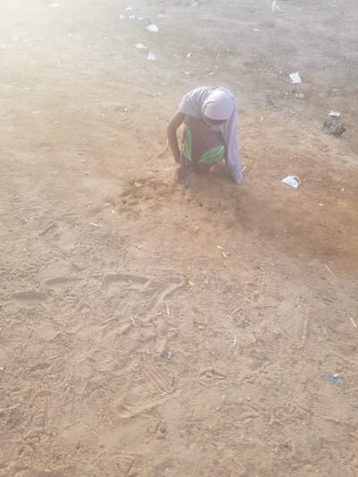 The image shows an individual in Dabawa, Nigeria, squatting on a sandy, dirt-covered surface. The person is wearing a light-colored head covering, a patterned garment, and long sleeves. Debris, including small pieces of paper and other unidentified objects, are scattered around the surface. The environment appears to be an outdoor, possibly undeveloped or unpaved, area within the city. The individual appears to be interacting with something on the ground, but the specific action or object is not clearly discernible due to the angle and distance of the shot.