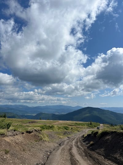 This is a full-frame, eye-level, outdoor, vertical shot of a mountainous landscape in Chorna Tysa, Ukraine. The foreground is dominated by a dirt road with tire tracks, which curves from the bottom right of the frame into the middle distance. The road is bordered by uneven, grassy terrain and some exposed soil and rocks on the right. In the mid-ground, there are rolling green hills with sparse trees and a few off-road vehicles visible on the road and the surrounding fields. The background features a series of layered, dark green mountains receding into the distance, with a hazy, blue-toned sky. The sky itself is partly cloudy, with large, dramatic white and gray clouds interspersed with patches of bright blue sky. The lighting suggests it is daytime, with the sun casting shadows and illuminating the landscape. There are no identifiable people visible in the image.