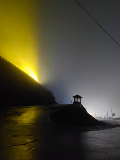 The image captures a dimly lit, atmospheric scene at night, likely in or near Kulang, India. A prominent feature is a large, dark wall illuminated from the left by a bright, yellow light that casts a radiant glow and creates a misty diffusion effect. The wall appears to be on an incline, and the road in front of it is wet and reflects the yellow light.
To the right, on a small, dark mound, stands a small, open-sided structure resembling a guardhouse or a small shelter, silhouetted against the hazy night sky. In the far distance, faint lights and structures can be discerned through the mist, suggesting a more developed area or possibly industrial elements. A power line or cable stretches diagonally across the upper right portion of the image. The overall impression is one of a remote, perhaps mountainous or elevated location, shrouded in fog and illuminated by artificial light, creating a surreal and somewhat mysterious ambiance. There are no visible people or animals in the scene.