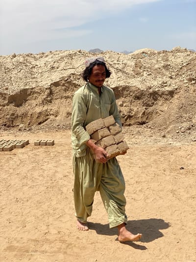 A man stands barefoot in a dusty, arid landscape in Deh-E Bagh, Afghanistan, carrying a stack of mud bricks. He wears traditional Afghan clothing: a light green tunic and loose pants, topped with a white and embroidered cap. The background features a large pile of dirt or mud, possibly where the bricks are made, and other dried mud bricks laid out. The sky is clear and bright, suggesting a hot, sunny day.
