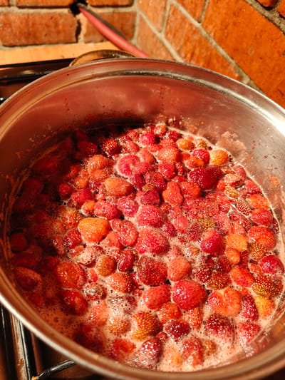 A close-up, overhead shot shows a stainless steel pot filled with boiling strawberries and their juices. The strawberries are a mix of red and orange hues, with some appearing plump and others slightly shriveled. They are bubbling and frothing, indicating they are actively cooking. The pot is resting on a black stovetop, partially visible at the bottom of the frame. In the background, a brick wall with a warm orange-red tone is present, suggesting an indoor setting. A red hose or pipe is also visible against the brick. The scene appears to be set in a kitchen, possibly in Starosemeikino, Russia, as this is a common domestic activity there, like making jam or compote.