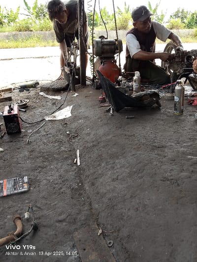 The image shows two men working on a motorcycle in a garage. The men are both wearing casual clothes and are bent over the motorcycle, which is partially disassembled. There is a compressor, tools, and a variety of other objects scattered around the garage. The garage is located in Kecamatan Anjatan, Indonesia, which is located at the coordinates -6.413, 107.953. The floor of the garage is made of concrete and is dirty. There is a piece of plastic sheeting on the ground next to the motorcycle. The men appear to be focused on their work and are interacting with each other. The environment is messy and cluttered. The time is 14:05 on April 13, 2025.