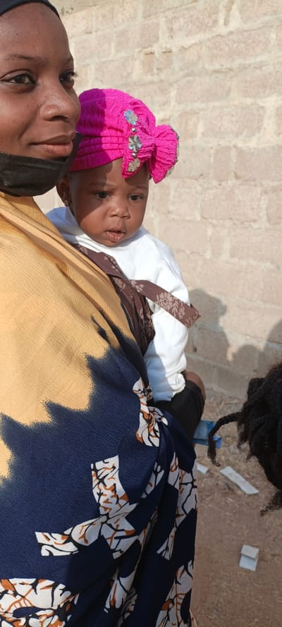A woman wearing a black mask holds a baby in a carrier. The baby wears a bright pink floral-embellished head covering. The woman wears a yellow and blue patterned garment. The baby has a slight smudge of something near their mouth. Background shows a light brown brick wall and dirt ground with debris. A person with dark dreadlocks stands on the right side of the image.