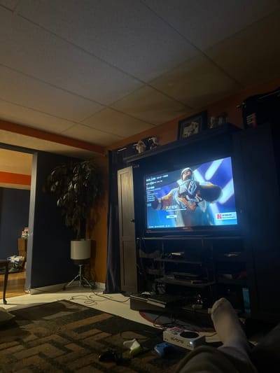 A television displays a hockey game. Below it is a cabinet with a dark curtain obscuring the bottom. To the right is a gaming console, controllers, and a white athletic sock. A faux plant stands in a white pot near a dark blue wall. A rug with a geometric pattern lies on the floor. The room appears cluttered with wires. The ceiling is textured and has a border along the wall.