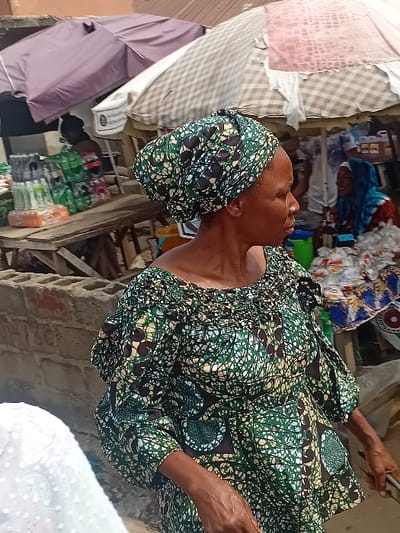 An older woman wearing a green and black patterned dress and headwrap is seen from the chest up, holding what appears to be a cellphone in her right hand. The setting looks to be inside a cluttered building, with fabrics piled in the background. A light pink umbrella and stacked packages are visible in the lower-left corner. The image has been rotated counter-clockwise, causing a tilted orientation.