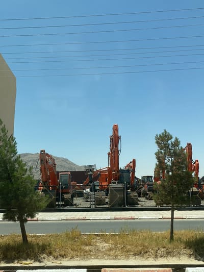 This image depicts an outdoor scene in Shurandam, Afghanistan, featuring a large collection of heavy construction machinery under a clear sky. In the upper portion, a bright blue sky dominates, traversed by five parallel, dark horizontal power lines. A light-colored building or wall is partially visible along the left edge of the frame.
The foreground consists of a paved road with a distinct red and white curb marking its edge, separating it from an unpaved median strip of dry, light-brown grass and scattered greener vegetation. Two evergreen trees, appearing pine-like, stand in this median strip, partially obscuring the view.
Behind the road and trees, a fenced enclosure holds numerous large orange excavators, many identifiable as Hitachi brand, parked closely together. Their booms and buckets are oriented vertically or at various angles, suggesting they are not in active operation. The dark metal fence encloses the machinery. In the background, beyond the machinery and enclosure, hazy light-brown mountains rise under the bright sky. No individuals are visible in the scene.