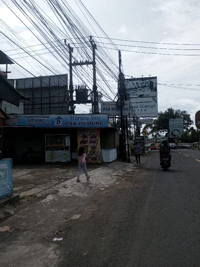 A street scene features "Warung Sate Pak Dudung," a food stall advertising "Sate Kambing," "Sate Ayam," and "Sate Maranggi." A young girl walks past the stall on the sidewalk. Further down the road, a man sweeps while a person on a motorbike drives by. Signs for "SUNDANESE RESTO Ban Wangi BISTRO" and "BRASCO COLES" are visible. Overhead, a complex network of utility wires crisscrosses the sky. Billboards and other signage adorn the scene, contributing to the bustling atmosphere.