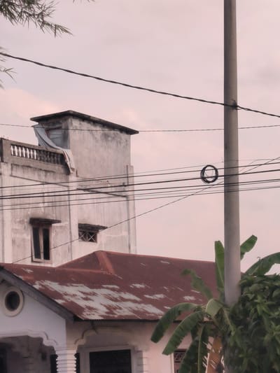 The image shows a street scene in Blangbitra, Indonesia. In the foreground, a weathered utility pole stands amidst lush greenery, including banana leaves. Overhead, a tangle of power lines crisscrosses the sky, one line featuring a coil of cable. The background reveals two buildings: a two-story white building with architectural details like a round window and a rusty red roof, and a taller, dilapidated white building with a balcony. The sky is overcast with a pink hue.