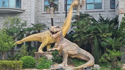 The image shows two dinosaur sculptures in a garden setting in Ulsan, South Korea. A Tyrannosaurus Rex model is in the foreground with its mouth open, and a Brachiosaurus model stands behind it. Palm trees, bushes, and other greenery surround the dinosaurs. A large, multi-story building constructed of light-colored stone is visible in the background.