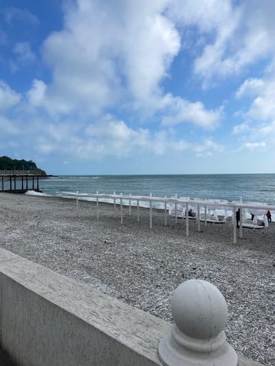 The image shows a pebble beach on a cloudy day in Olginka, Russia. White beach canopies and lounge chairs line the shore. A concrete wall with a decorative sphere is in the foreground. A wooden pier extends into the water. The sky is blue with white clouds. The sea is a dark grey-blue.