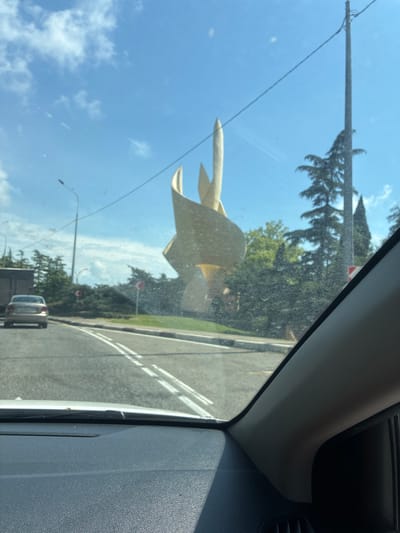 The image shows a view from inside a car, looking out through the windshield at a roundabout in Novomikhaylovsky, Russia. A large, golden abstract sculpture is visible in the center of the roundabout, which is surrounded by greenery and trees. A silver car is driving on the road leading into the roundabout, and a traffic light is visible in the distance. The sky is blue with some clouds. The windshield appears to be dirty.