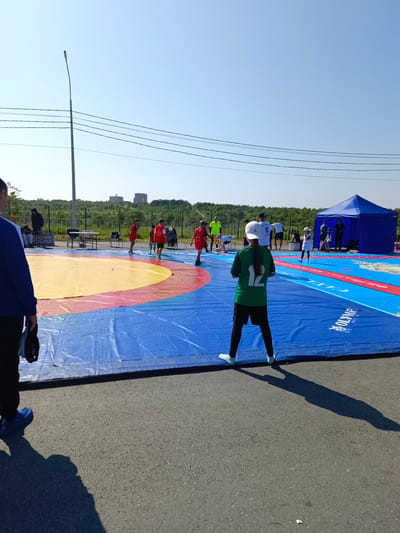 An outdoor wrestling event is taking place in Korsakov, Russia, on a bright, sunny day. The central focus is a large, blue wrestling mat with concentric red and yellow circles in the center, laid out on asphalt. Several individuals, mostly children, are present on and around the mat, some wearing red athletic tops, others in green and white. One child in a green shirt with the number "12" on the back is facing away from the camera, standing on the blue portion of the mat. A blue tent, likely for shade or storage, is situated to the right of the wrestling area. In the background, a line of trees borders a gentle hill, and several modern buildings are visible in the distance. A street lamp and power lines are prominent in the sky. A man wearing a blue jacket and black pants, holding a dark object, is in the foreground on the left, casting a long shadow. Other adults, possibly coaches or organizers, are also present. The overall scene depicts an organized sports activity in an urban park or open area.