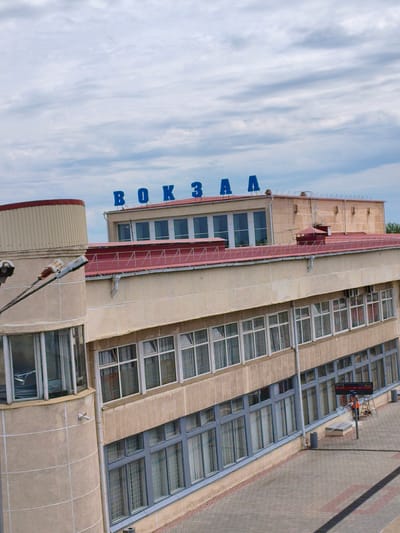 The image shows a street-level view of a beige, multi-story building with a red roof. The building has a curved section on the left with a red-capped cylindrical structure on top. Blue letters spelling "ВОКЗАЛ" (Russian for "station") are visible on the upper part of the building. The building features numerous windows with white frames on the upper floors and darker frames on the lower floors. A streetlamp with two lights is visible in the foreground. The ground in front of the building is paved with interlocking stones and features a dark band running diagonally. To the right, a digital display board showing "51:09" and "23:00" is mounted on a pole, with a person in an orange vest standing on a ladder next to it. The sky is overcast with a cloudy, light blue hue. The overall scene depicts a train station or similar public building in Buzuluk, Russia.