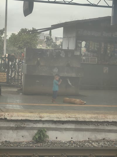 The image depicts a railway platform in Chhanera, India. A young child wearing a blue t-shirt and dark shorts stands near a concrete structure, holding what appears to be a mobile phone or small camera. A light brown dog is lying down on the platform in front of the child. In the background, there is a stall with various items displayed, possibly snacks or drinks. Behind the stall, trees and buildings are visible. The platform shows signs of wear, with some cracked concrete and debris. The sky is overcast.