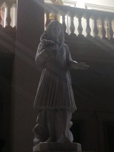 A stone statue of a woman holding a bouquet of flowers stands in a dimly lit indoor space. The statue is dressed in a long, flowing robe with intricate pleats. Her hair is styled in loose waves, and she has a serene expression on her face. Her right arm is extended, and her palm is open, as if offering something. The background features a balustrade with ornate carvings, illuminated by rays of light filtering from an unseen source. The overall atmosphere is solemn and reverent. The statue is likely a religious or historical monument, possibly located in a church, museum, or historical site. The mention of Xuân Hương - Đà Lạt, Vietnam suggests this is a specific location within the city.