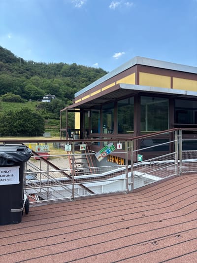 This is a medium-shot, eye-level, outdoor, daytime photograph taken in Lehmen, Germany. The foreground features a brown, textured surface with dark lines, possibly a deck or walkway. A black trash bin with a black lid and a white label that reads "ONLY! CARTON & PAPER!!!" is on the left. In the midground, a metal railing with a net barrier separates the walkway from a lower level. A set of stairs leads down, with several signs visible on the wall. One sign reads "LEHMEN", another has a picture of a medical cross, and a third sign shows a person running with a green arrow. Beyond the railing and stairs is a modern building with large windows and a yellow exterior. The building is situated on a hillside covered in lush green trees and vegetation. The sky above is blue with a few scattered white clouds.
