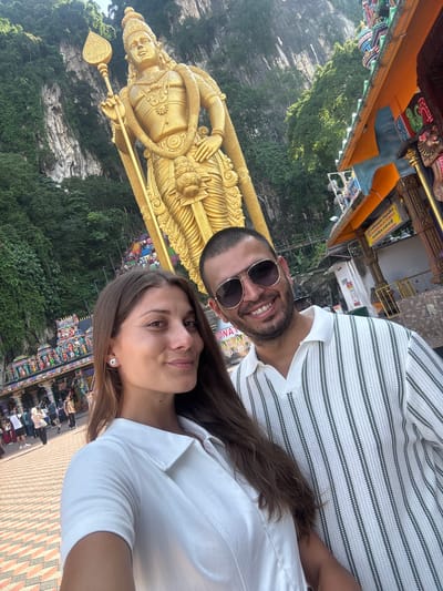 A man and a woman are posing for a selfie in front of a large, golden statue of a Hindu deity at the Batu Caves in Selayang Municipal Council, Malaysia. The man is wearing sunglasses and a striped shirt, while the woman is wearing a white shirt. In the background, there is a cliff face covered in greenery and a colorful temple structure. People are seen walking around in the background as well.