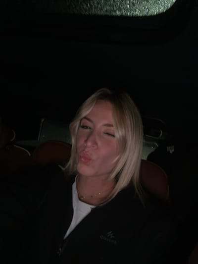 A young woman with short blonde hair is seated, possibly in a car at night, visible through the rain-streaked window on the left. She is turned towards the viewer, giving a kissy face, and has one eye squinted shut. She wears a black jacket over a white shirt, and a delicate gold necklace is visible. The background is dark, with hints of a car seat and a headrest behind her.