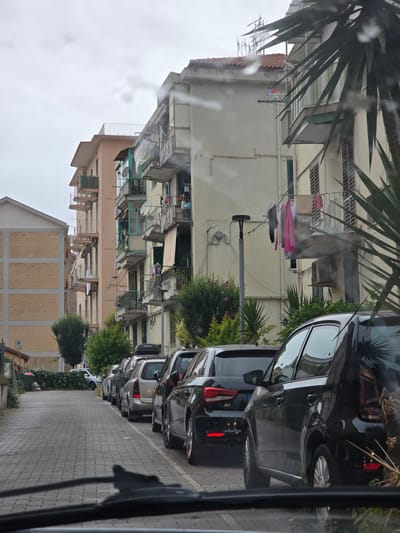 The image captures a street scene in Salerno, Italy, likely in a residential area characterized by apartment buildings and parked cars. The perspective is from inside a vehicle, looking out through the windshield, which shows streaks of water, suggesting it might be raining or the windshield is wet.

Several cars are parked in a line along the cobblestone street. The buildings are multi-story, with balconies where laundry is hanging out to dry. Some buildings appear to have air conditioning units mounted outside. Vegetation, including trees and bushes, is visible alongside the street and between the buildings. A street light stands on the right side of the street. A small sign with a parking symbol is visible on the left side of the street. The sky is overcast. The scene depicts an ordinary urban environment in Italy.
