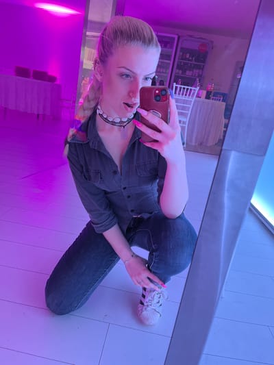 A young woman is taking a selfie in front of a mirror. She is kneeling, wearing jeans, a dark button-down shirt, and white Adidas sneakers. She has pink nail polish and is holding a phone with a maroon case. The reflection in the mirror shows a display of items behind her, possibly a refrigerator and some shelving units. The overall lighting is a mix of blue and purple hues. The location is likely indoors, in a commercial setting in Rakovski, Bulgaria.