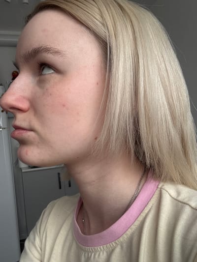A side profile shot of a person with blonde hair, light skin, and light eyebrows is presented. The person is wearing a light-colored shirt with a pink trim around the neckline. A silver necklace is visible around their neck. The background is blurred, but suggests an indoor setting. There is no information in the image that specifies the location is in the city of Novoe Devyatkino, Russia.