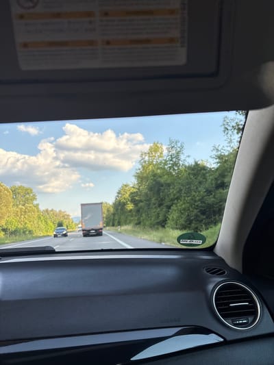 The image is taken from the interior of a car, looking out through the windshield. The view is of a highway with trees lining the sides. A gray truck is in the right lane, and a dark car is in the left lane, both moving away from the camera. The sky is blue with white clouds. The dashboard of the car is visible in the foreground, with an "AIRBAG" label and an air vent. A circular green sticker with white lettering is visible on the right side of the windshield. This sticker has the text "BAD VÖCK" in white, which is an abbreviation for Bad Vöcklin, a town in Germany. The image is likely taken in the city of Iffezheim, Germany, given the context of the sticker.