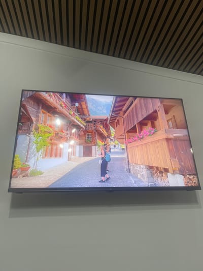 The media file shows a television screen displaying an image. The image on the screen depicts a charming, narrow street in a European village, likely in the Alps, given the traditional wooden architecture and the stacked firewood visible on the right. The buildings are made of dark wood with balconies overflowing with vibrant pink and red flowers. In the foreground of the screen, a person is visible walking away from the camera, wearing a backpack. The street itself is paved with cobblestones. The scene suggests a pleasant day with ample sunlight casting shadows. The overall impression is one of a picturesque, historic, and tranquil location. The television is mounted on a light-colored wall, with a textured, horizontally striped wall to the left, partially visible. The image on the screen is oriented vertically, suggesting the original photo was taken in portrait mode or the screen is rotated. No visible text is present in the media file.
