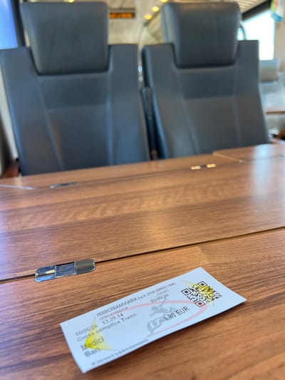 The image shows a train ticket on a wooden table in a train carriage. The ticket is white with some red and yellow accents. It features a QR code, a date "10/06/26", a price "1.80 EUR", and text including "FERROTRANVIAria S.p.A.", "data emissione", "validità", "Corsa semplice Treno", "Medici", and "Bari". The background shows the backs of two dark gray headrests of seats in a train. The overall environment appears to be a passenger train compartment. The presence of "Bari" on the ticket suggests a journey related to or originating from the city of Bari, Italy.