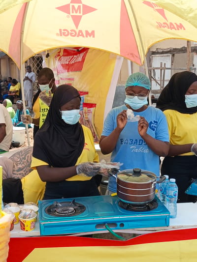 In Tarauni, Nigeria, a yellow and red tent provides shade for an outdoor event, possibly a food demonstration or market stall. Three individuals are the primary focus, all wearing face masks. Two women, dressed in yellow shirts and headscarves, are preparing food. The woman on the right, wearing a black headscarf, is holding a plastic bag and appears to be handling ingredients. To her left, another woman in a black headscarf and yellow shirt is also preparing food, wearing gloves. In the center, a third person, also wearing a face mask and a blue hairnet, is holding a small plastic bag with what appears to be food contents, looking at it intently. This individual wears a blue t-shirt with text that reads "PP| FABADOTables".

In front of the people, a blue portable gas stove is set up with a brown pot on top. Several small containers, likely for food, are arranged to the left of the stove. Two bottles of water, one labeled "Pure Life", are to the right of the pot. The scene is brightly lit, suggesting it is daytime. In the background, other people are visible, some sitting and others standing, hinting at a gathering or public event. The tent itself has a large logo on it, a red star with a white "M" inside, and text that reads "MAGGI" in reverse. The overall atmosphere seems to be one of activity and preparation for serving food.