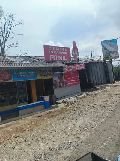 A roadside stall with a corrugated metal roof displays signs for "Mie Ayam & Es Campur FITMIL," "Teh Pucuk Harum," and "Minimania 2.3 km Lagi." Advertisements for "Juara" and "Aroma Manis Juara Apel" are visible on the storefront, along with another promoting "Juaraka Hidupmu Juara." Merchandise lines the shelves behind the blue-trimmed counter. The ground is uneven and appears to be gravel. Trees and a partly cloudy sky are in the background.