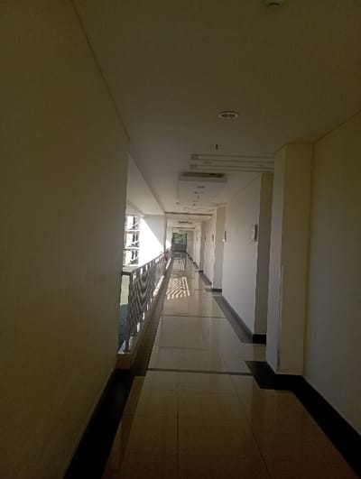 The image depicts a long, unoccupied indoor-outdoor corridor within a modern building in Batulawang, Indonesia. The perspective is a straight-on view down the length of the hallway. On the left, an open-air section features a silver-colored metal railing, allowing natural light to enter and revealing parts of adjacent building structures beyond. The right side is defined by a solid light-colored wall, punctuated by evenly spaced columns or alcoves, with several rectangular wall-mounted objects visible further down. The floor is composed of large, reflective light-colored tiles with darker brown or black rectangular borders running down the center and along the base of the right wall, showing clear reflections of the ceiling and light sources. The ceiling is flat and light-colored, fitted with multiple recessed circular lights and rectangular ventilation or decorative panels. A smoke detector is visible on the ceiling in the foreground. Sunlight casts distinct diagonal shadows across the left portion of the floor. No individuals, actions, or interactions are present in the scene. The environment appears clean and well-maintained.