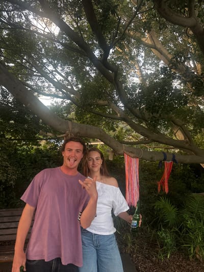 This image, captured outdoors in Thirroul, Australia, depicts two individuals standing under a large tree with dense green foliage.

On the left, a male with light brown, curly hair wears a loose-fitting purple short-sleeved t-shirt and dark trousers. He faces the camera directly, mouth open with his tongue extended, and his right hand performs a "shaka" gesture. His left hand is partially visible at the bottom left, appearing to hold an object.

On the right, a female with long, light brown hair wears a white off-the-shoulder top and light blue wide-leg jeans. She stands next to the male, smiling subtly at the camera, and holds a green glass bottle, identified as a Hahn beer bottle by its label, in her left hand.

The background features the prominent branches and leaves of the large tree, with sunlight filtering through the canopy. One lower tree branch is decorated with colorful fabric streamers: a long vertical arrangement of pink, orange, and white strands hangs to the left of the female, and shorter red and orange strands tied with dark blue fabric are further to the right. Dense green vegetation and plants are visible behind the individuals and to the right. A wooden bench is partially visible behind the male on the left. A distant hill is discernible through the upper tree branches.