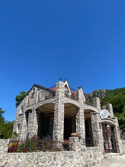 The image depicts a stone building in Sotonići, Montenegro, under a clear blue sky. The structure has multiple levels with arched openings and wrought iron railings. A satellite dish is mounted on the upper level. The building is constructed from roughly cut stones of varying sizes. The surrounding environment includes greenery and a hillside.