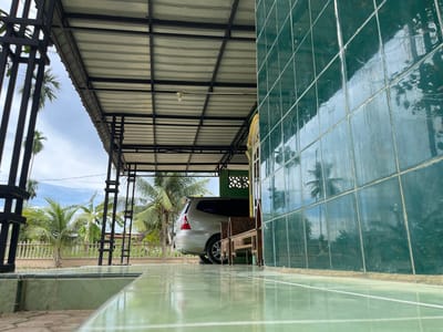 The image depicts an outdoor scene under a metal roof supported by black metal pillars. The foreground is dominated by a glossy green tiled floor. To the right, a wall covered in large, dark green tiles reflects the surroundings. In the midground, a silver minivan is parked under the covered area. Adjacent to the minivan is a wooden bench with intricate carvings. Beyond the covered area, palm trees and other greenery are visible, suggesting a tropical or subtropical environment. A light blue sky with scattered clouds is visible in the background. The overall impression is one of a covered outdoor space, likely a driveway or patio, attached to a building with a tiled facade. The scene is located in Benuaraja, Indonesia.