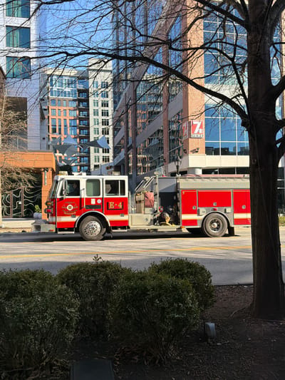 A red fire truck is parked on a street next to a sidewalk with green bushes. The truck has white accents and the number "E-4" displayed. In the background, there's a large building complex with reflective glass windows and brick accents. A news channel logo "7 NEWS" is visible on one of the buildings. Bare branches from a tree hang in the foreground, framing the scene.