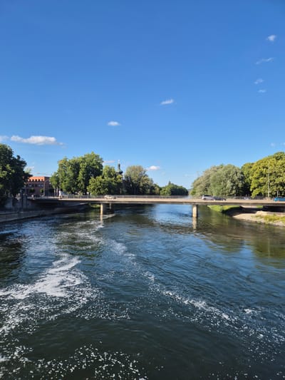 The image depicts a wide river flowing through the city of Landshut, Germany. A concrete bridge spans the river, carrying vehicular and pedestrian traffic. On the left bank, a multi-story brick building is visible, partially obscured by trees. A church spire with a green dome rises above the trees in the background. The right bank is lined with lush green trees. The river's surface is a mixture of dark water and frothy white foam, indicating a current. The sky is a clear, vibrant blue with a few scattered white clouds. The lighting suggests it is a sunny day, likely late morning or early afternoon. There are several cars and a few people visible on the bridge.