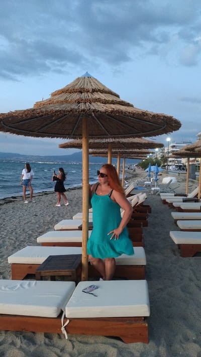 An eye-level, full shot shows a woman with long, reddish-orange hair and sunglasses standing on a sandy beach under a thatched umbrella, posing with her hand on her hip and the other on the umbrella's pole. She is wearing a teal, sleeveless dress with a ruffled hem. Behind her, several rows of empty lounge chairs with white cushions are arranged. In the background, the sea is visible with waves and distant hills. To the left, two younger women are standing near the water; one in a white t-shirt and denim shorts, the other in a black dress, holding a phone. The sky is overcast with muted blue and gray clouds. The scene appears to be a resort or public beach area in Neoi Epivates, Greece, at dusk or during an overcast day.