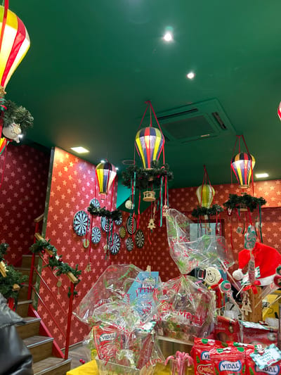 This indoor image, taken in Chalkida, Greece, captures a vibrant and festive retail or event space adorned with colorful decorations. The scene is brightly lit by artificial lights, reflecting on a smooth, solid green ceiling where an air conditioning unit is visible.
Hanging from the green ceiling are several elaborate, hot air balloon-shaped lanterns or decorations. These balloons are striped in yellow, blue, red, and white, and each features a small "basket" structure below. Many are further embellished with green Christmas garlands and small festive ornaments, suggesting a holiday theme.
The floor or main display surface is covered in a striking red material patterned with white stars, enhancing the celebratory atmosphere. Arranged on this star-patterned surface are numerous circular, dartboard-like decorations, adding a playful element to the environment.
On the right side of the image, various merchandise items are displayed, predominantly wrapped in clear plastic. These appear to be sweets or gifts. Several red packages are prominently visible, featuring the brand name "VIDAL" and the text "Merry Christmas." One package displays a white Mickey Mouse head. A yellow price tag with "4.40€" indicates the cost of an item. A red Santa hat can be seen towards the top right, reinforcing the Christmas theme. A person's hand, partially obscured by a jacket, is visible in the bottom right corner.
Overall, the image depicts a cheerful, holiday-themed display, possibly a shop specializing in candies or festive goods, set in a European location as evidenced by the currency.