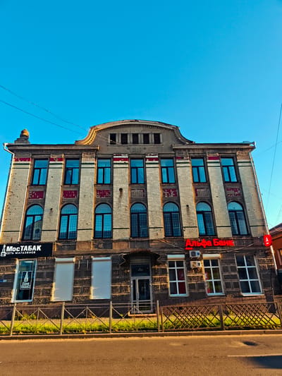 The image shows a wide, low-angle, exterior, daylight shot of a multi-story brick building. The building is located in Kimry, Russia. The facade of the building is adorned with decorative elements, including red and green stripes and carved stone details. There are numerous windows, many of which are arched. On the first floor, there are storefronts. A sign with Cyrillic text reads "МосТАБАК" and another sign with red Cyrillic text reads "Альфа Банк". A metal fence runs along the front of the building, with a strip of green grass and a sidewalk behind it. A paved road runs in front of the sidewalk. The sky is clear and blue. Electrical wires are visible against the sky.