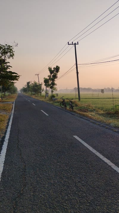 This image depicts a tranquil dawn scene on a paved road in Sumberejo, Indonesia. The road, marked with white lines, stretches into the distance, flanked by lush green rice fields on the right and trees and vegetation on the left. Two bicycles are parked by the side of the road, suggesting a pause in a journey. Utility poles with power lines are visible, indicating the presence of infrastructure. The sky is a soft gradient of pale orange and gray, with a hint of fog or mist settling over the landscape, creating a serene and peaceful atmosphere. The overall impression is one of quiet rural beauty at the beginning of a new day.