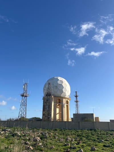 An outdoor daytime scene in Dingli, Malta, depicts a radar station under a clear blue sky with scattered white cumulus clouds. The central element is a large, white, geodesic radome situated atop a multi-story, cylindrical building constructed from beige masonry. To the left and right of this central structure are tall, metal lattice communication towers equipped with various antenna arrays. The entire facility is enclosed by a beige perimeter wall, topped with razor wire, indicating a secure area. The foreground consists of an uneven, rocky, green grassy field, with numerous light-colored stones scattered across the terrain and sparse vegetation. No individuals or active interactions are visible within the frame.