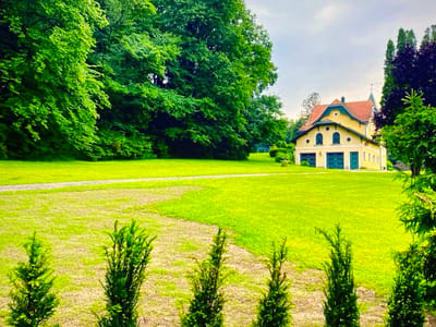 The image depicts a charming yellow house with a red roof situated in a lush green landscape. The house features arched windows and two large garage doors. It is surrounded by manicured lawns and mature trees, with a pathway leading across the expansive green area. The scene suggests a tranquil setting within the city of Purkersdorf, Austria. The foreground is occupied by several evergreen shrubs, adding depth and texture to the composition. The sky above is overcast.