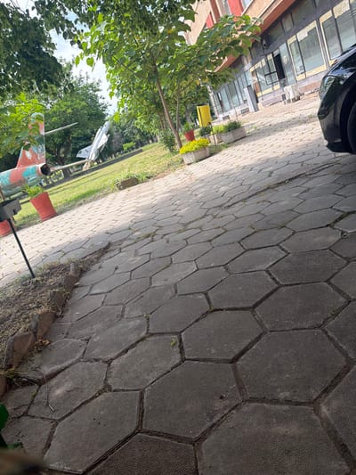 The image shows a paved walkway with hexagonal pavers, with greenery and trees to the left. In the background, partially obscured by trees, is a white jet plane on display. A painted aircraft tail with red and green accents is visible in the foreground, near a pole. The photograph appears to be taken outdoors, likely in a park or memorial area. Based on the prompt, this could be in Dolna Mitropolia, Bulgaria.