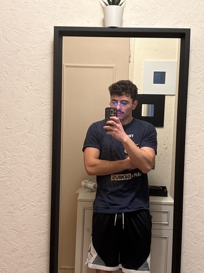 This image depicts a mirror selfie taken indoors in Rennes, France. A male individual, appearing to be an adult, is visible from the waist up, reflected in a large, black-framed mirror. He has dark, curly hair, a mustache, and is wearing round spectacles that reflect blue light. He is dressed in a dark blue short-sleeved t-shirt with reversed text (including "GENIUS") and black athletic shorts featuring white and grey panels with a visible Jordan logo. His arms are crossed, and he is holding a dark-colored smartphone with a textured case in his right hand, positioned to capture his reflection.
The environment reflected in the mirror includes a light-colored closed door to the left of the individual. Below his waist, a white vanity or chest of drawers is visible, with a black tray and other indistinct items on its surface. Above the vanity, two pieces of wall art are displayed: a white square frame containing a dark blue square, and below it, a black square frame with multiple light blue vertical stripes. A small white potted succulent rests on the top edge of the mirror frame. The wall surrounding the mirror is textured with a light, off-white finish.