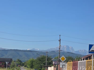 The image depicts a clear, blue sky above a rural scene in Ir, Russia. In the foreground, a road winds through a small settlement with houses and a colorful fence. Power lines crisscross the sky, supported by utility poles. Behind the houses and trees, a range of lush green mountains forms a backdrop, with snow-capped peaks visible in the distance. The overall impression is one of a peaceful, sunny day in a mountainous region.