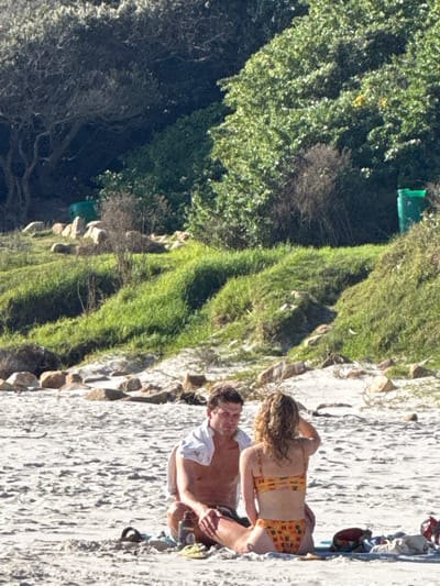 The image shows a couple on a beach next to a grassy slope. The man is wearing a white t-shirt and is sitting on the beach. The woman is wearing a patterned swimsuit and is kneeling next to him. They appear to be interacting, possibly talking or embracing. The beach is sandy and there are some rocks along the edge. The location appears to be Hout Bay, South Africa, based on the landscape and vegetation.