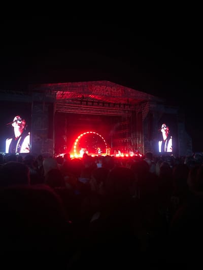 This image depicts a night-time outdoor music concert held in Ribamontán al Mar, Spain. In the foreground, a large crowd of spectators is visible as dark, undefined silhouettes, observing the performance. The midground features a prominent, multi-level concert stage constructed from metal trussing, illuminated primarily by intense red stage lighting. Two large, vertical LED screens flank the central performance area, each displaying a close-up, brightly lit image of a male performer singing into a microphone. On the main stage, multiple figures, likely musicians, are present. A distinctive semi-circular array of bright red lights forms an arch behind the performers, and additional spotlights are visible. A banner spanning the top of the stage structure clearly reads "SANTANDER MUSIC 2023". The background is uniformly dark, indicating a night sky.